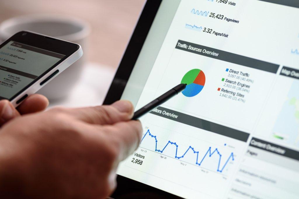 Una mano señala con un lápiz un panel de analítica con gráficos de visitas en una tableta, junto a un teléfono.