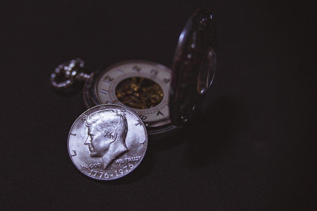 Una moneda junto a un reloj de bolsillo abierto sobre una superficie oscura.